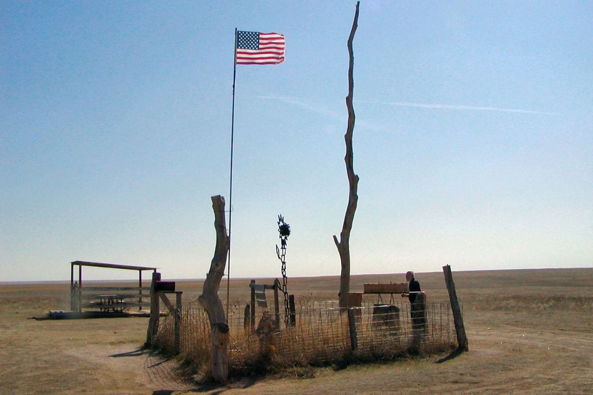 A picture of Mount Sunflower, the highest point in Kansas. Weathered sticks placed upright in a square with an American flag are in the picture. A man looking at the monument can be seen on the right.