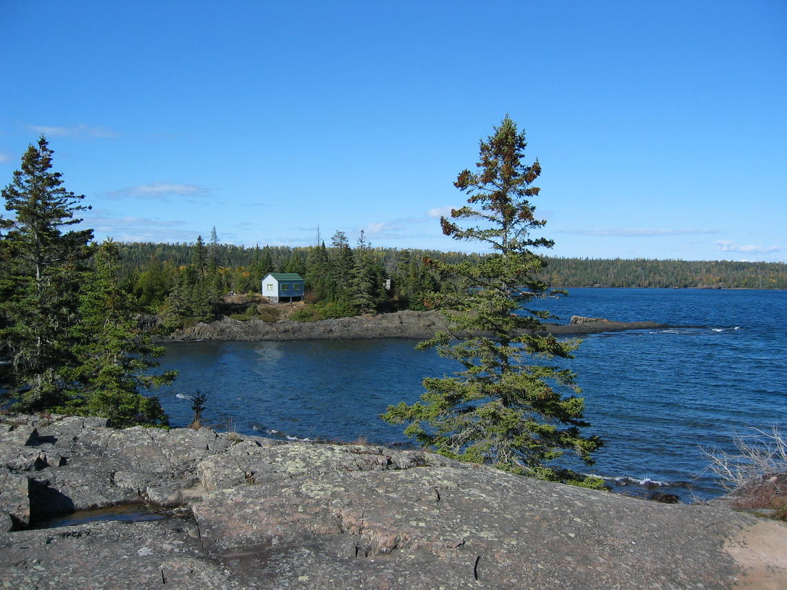 Photo of Isle Royale National park, Michigan, across an inlet, with rocky headland, blue lake water, and cabin on the opposite shore.