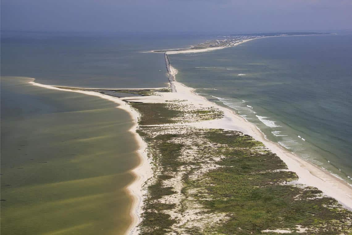 An aerial view of Dauphin Island, Alabama, shows a thin strip of road partially covered by sand, with undeveloped beach in the foreground and developed beach in the background. Photo: Karen Morgan, USGS. Public domain.